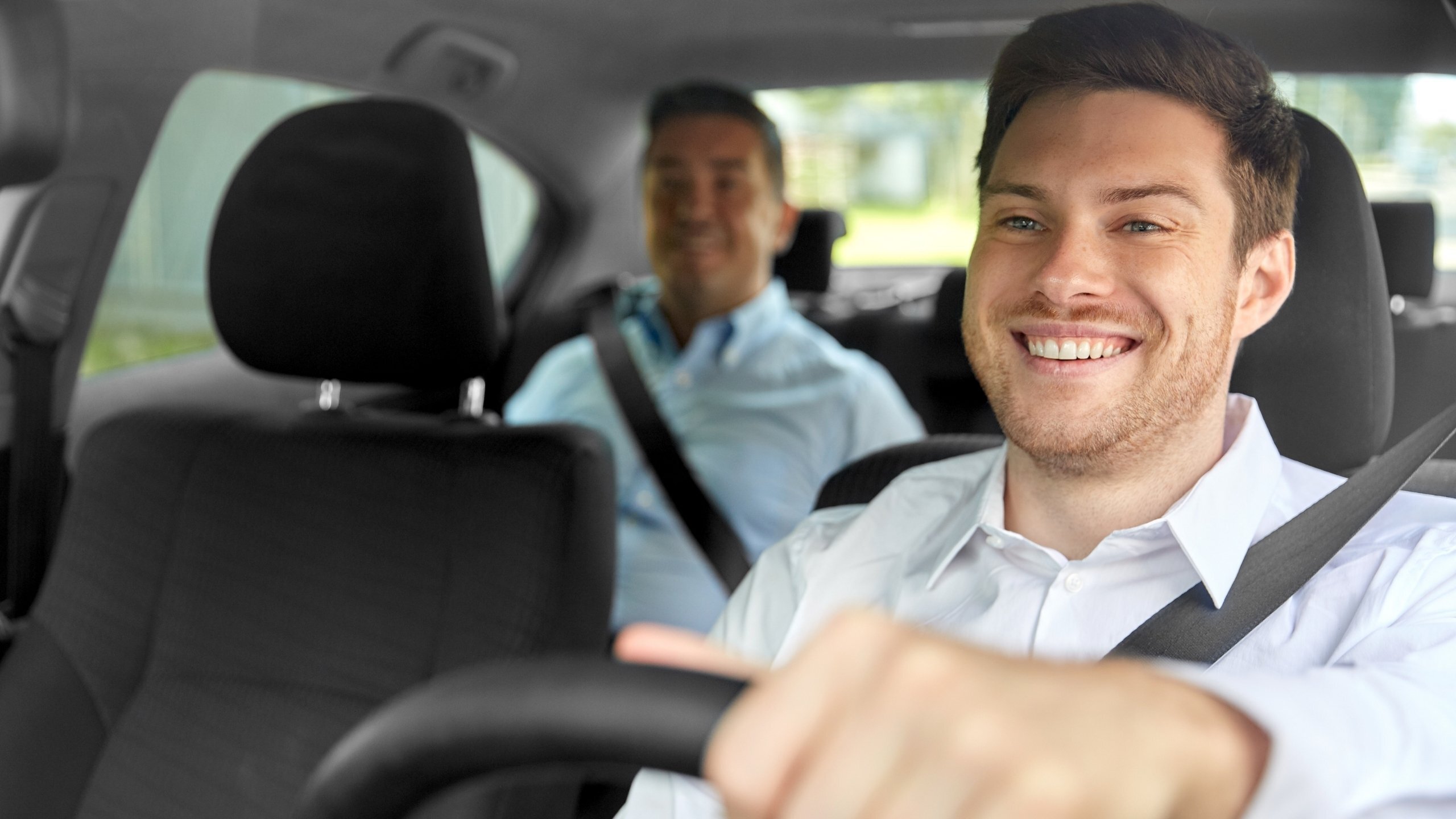 Adult learner driving with a smiling instructor sitting in the back seat, both wearing seatbelts inside a car.