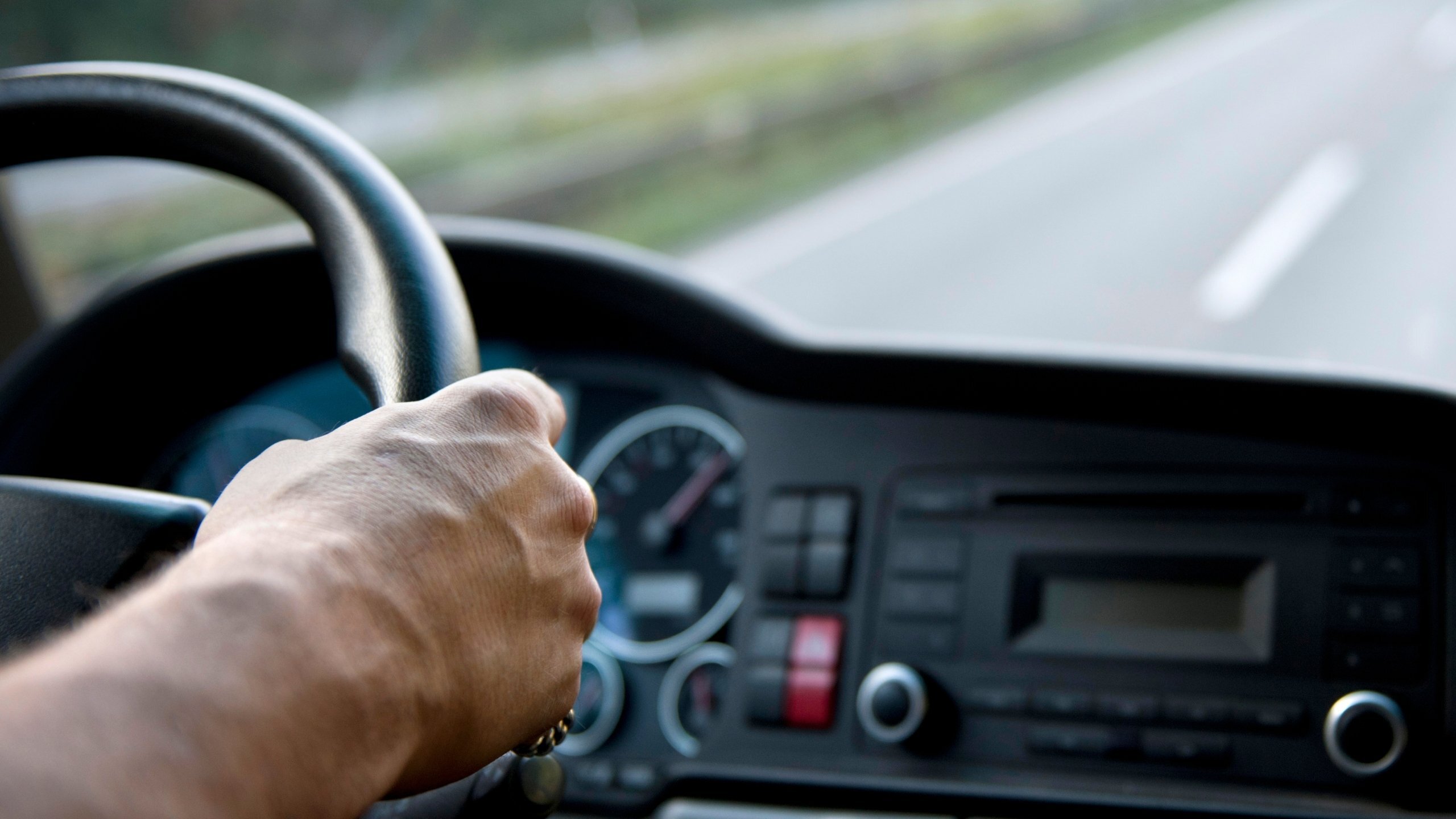Close-up view from the driver’s seat showing a person’s hand on the steering wheel while driving on an open road.
