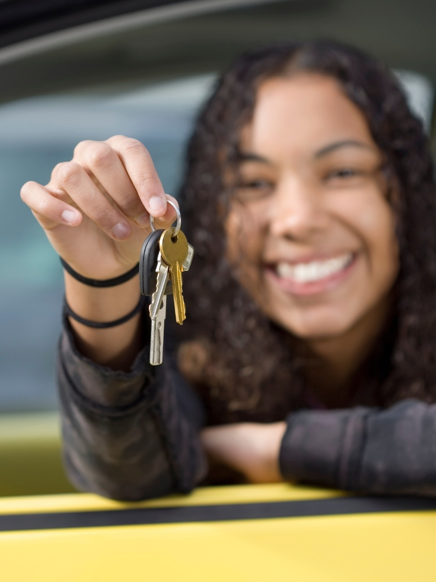 Smiling teen holding out a set of car keys from the driver’s seat of a yellow car.