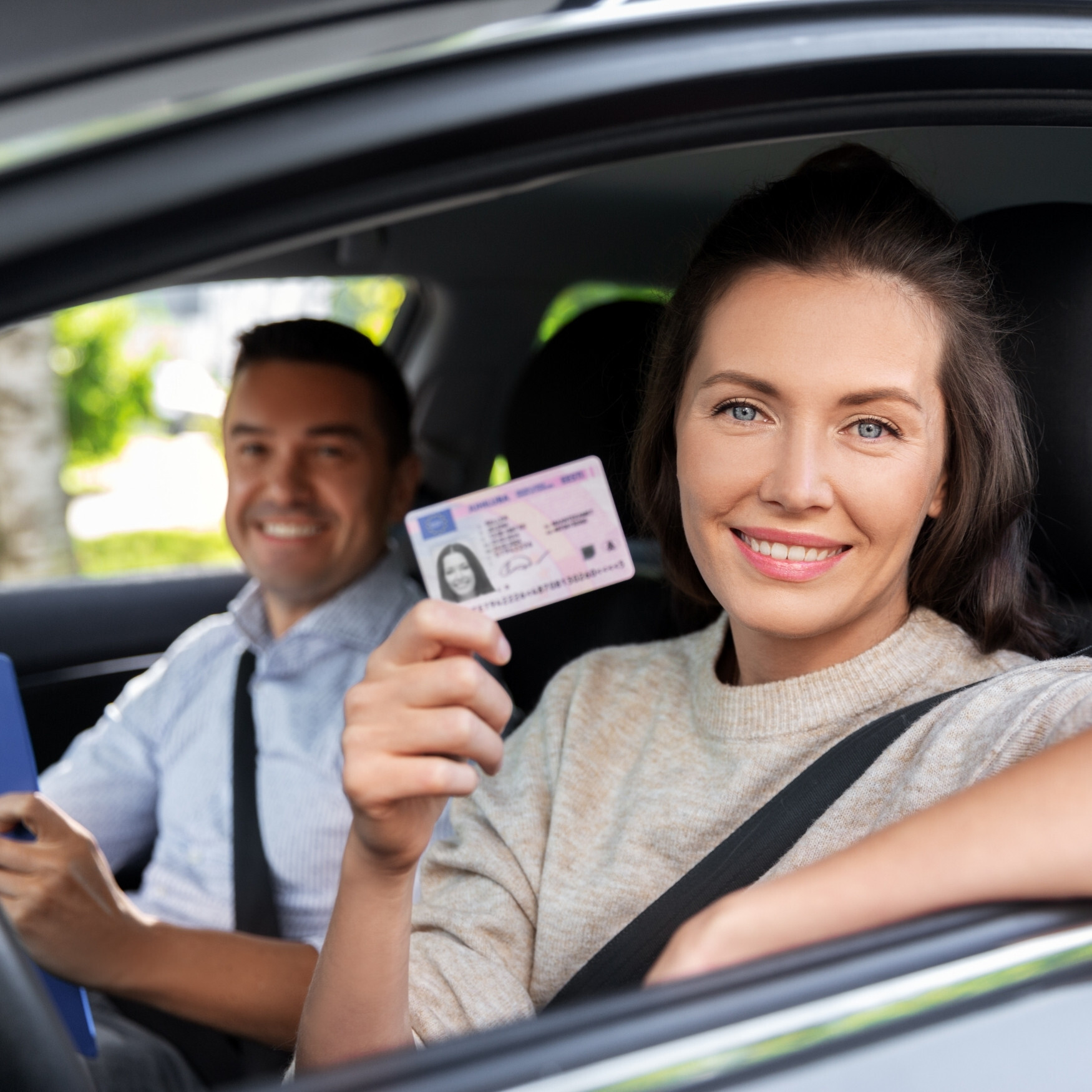 Woman sitting in the driver’s seat smiling and holding up her new driver’s license, with an instructor seated beside her.
