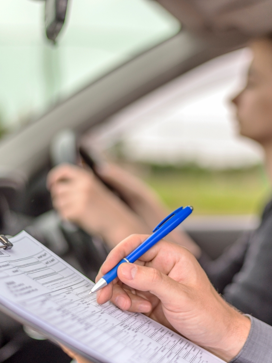 Driving instructor marking a checklist on a clipboard while a student drives during a road test.